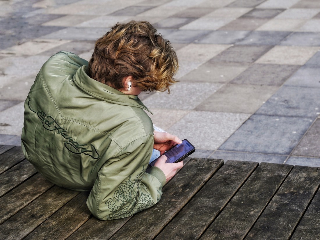 A teenager sitting alone on a bench, head bowed over a phone