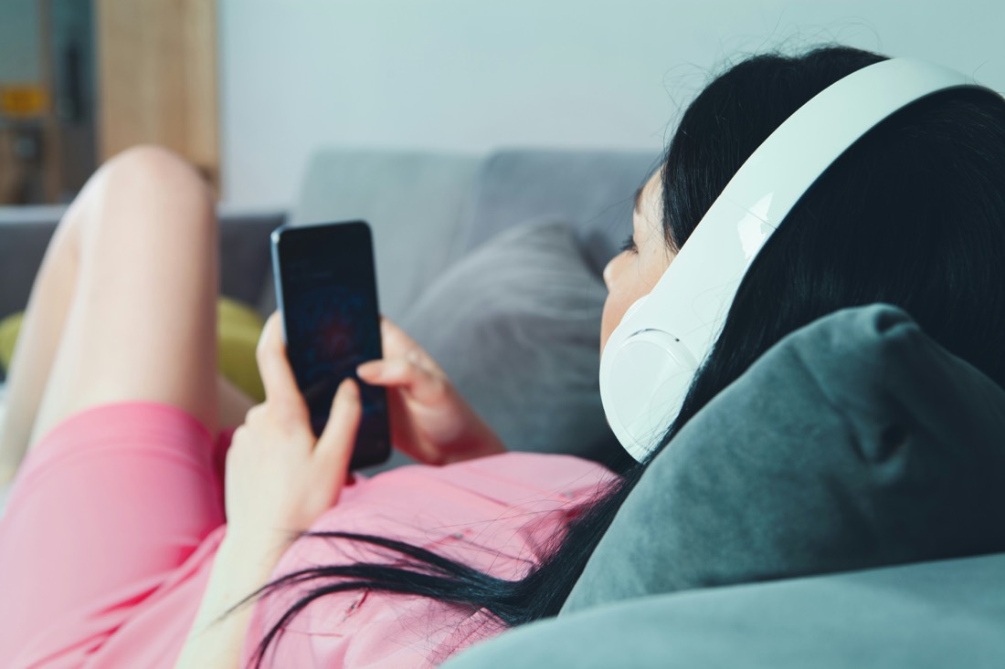 A young person lying on a couch with headphones, scrolling a phone